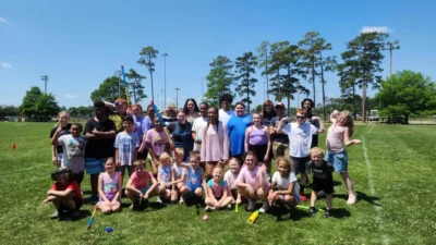 A group of smiling children and adults poses on a sunny day at a park, with green grass and tall trees in the background, conveying joy and camaraderie.