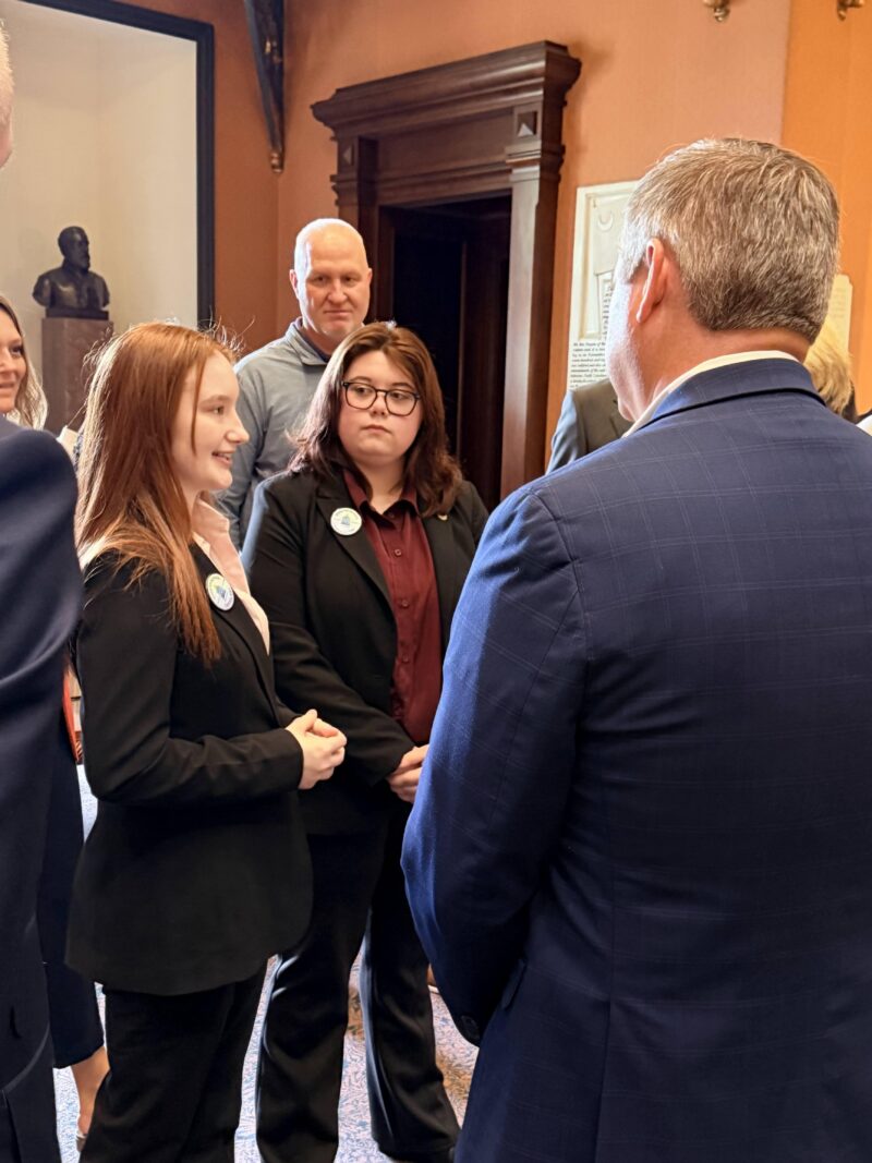 Two young women in suits engage in conversation with a man in a blue suit.