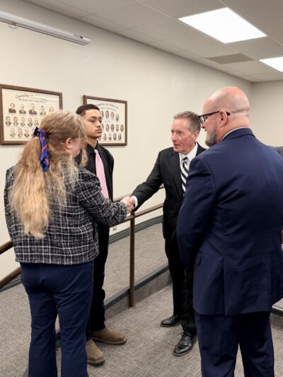 Four people in formal attire converse and shake hands in an office setting, with framed photos on the wall.