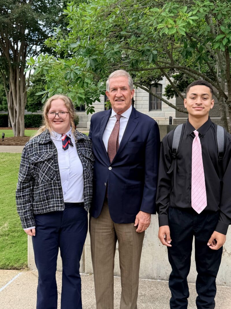 A man in a suit stands between two smiling students outside