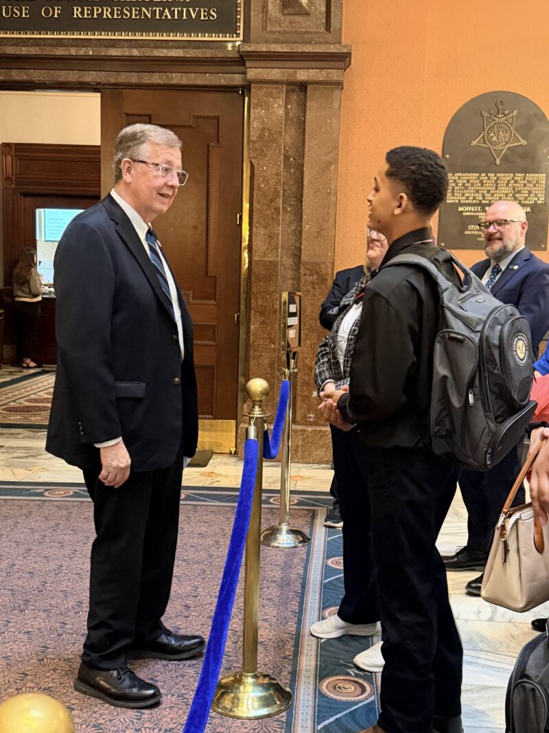 A man in a suit speaks with a younger man wearing a backpack, inside a government building