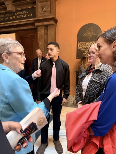 A woman in a blue suit converses with a group of young people in a formal setting