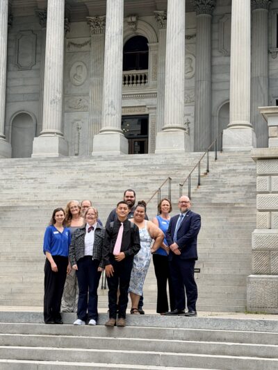 A diverse group of nine people stand smiling on the steps of a grand building